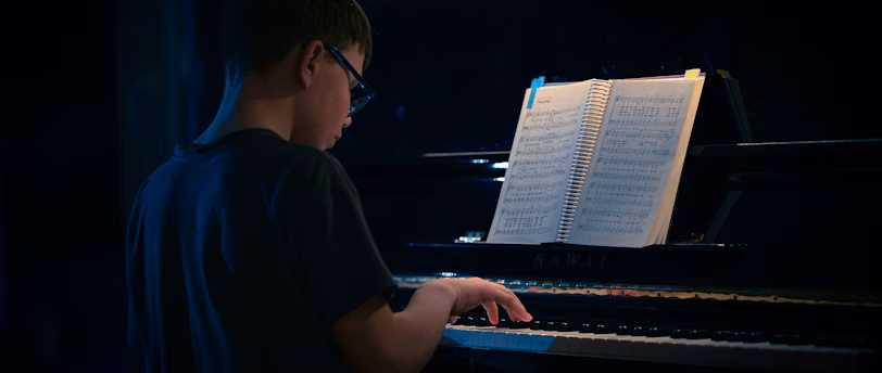 A young boy practising the piano in a dark room with a lit-up score