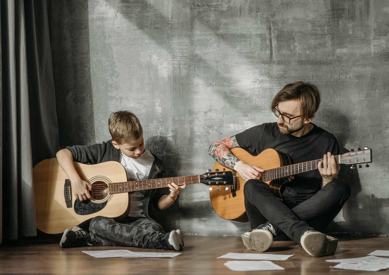 A guitar teacher and a young student sitting on the floor with their acoustic guitars, practicing together with sheet music spread around them.